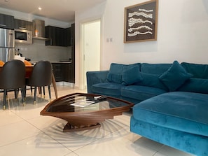 Una sala de estar moderna con un sofá azul, una mesa de centro de vidrio y una cocina al fondo.