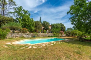 Una piscina rodeada por un muro de piedra, abundante vegetación y un edificio de piedra al fondo.