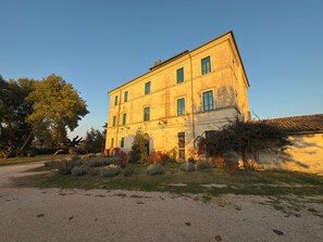 A two-story building with a symmetrical facade, multiple windows with green shutters, and a gravel driveway.