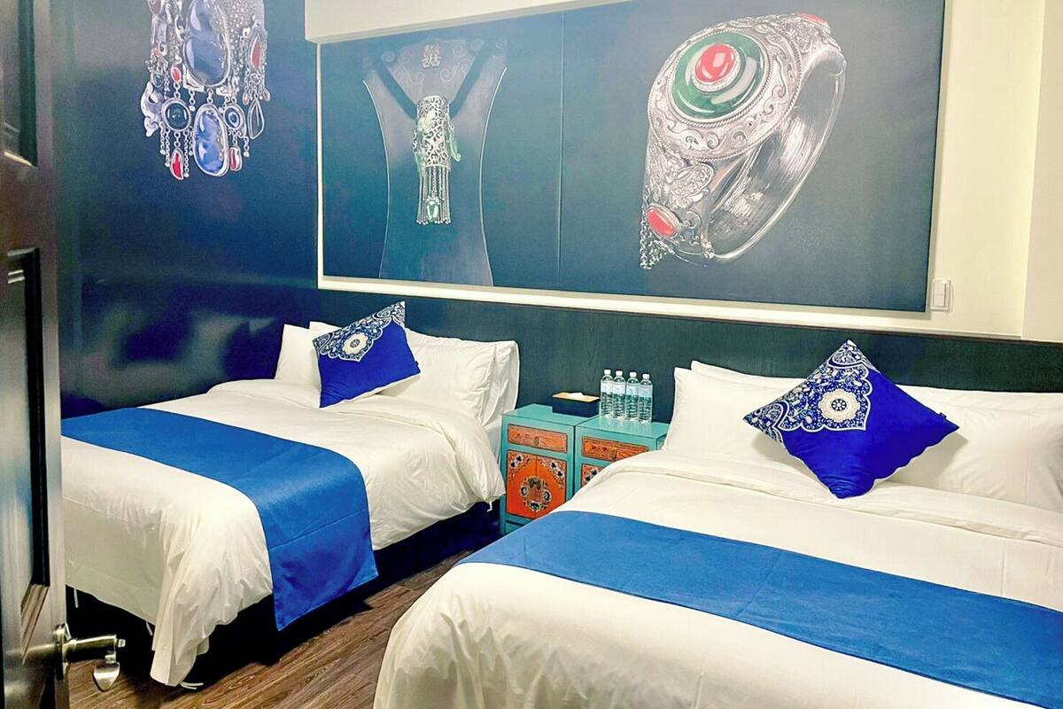 Habitación de hotel con dos camas, ropa de cama azul y blanca, y una pared decorada con joyas.