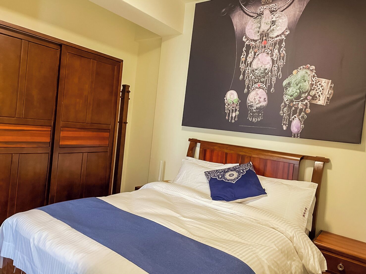 Een net opgemaakt bed met een blauw accentkussen, een houten garderobe en een groot poster van sieraden aan de muur.