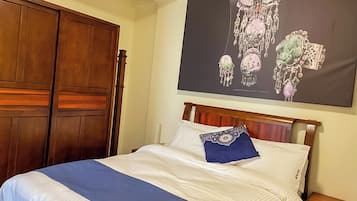 Een net opgemaakt bed met een blauw accentkussen, een houten garderobe en een groot poster van sieraden aan de muur.