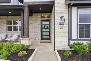 A building entrance with a brick facade, a door with a wreath, a welcome mat, and a number '300' on the wall. - LUXE COUNTRY FARMHOUSE with over 1 acre, dogs OK, 4bd, 3 ba fully furnished! (Gunter)
