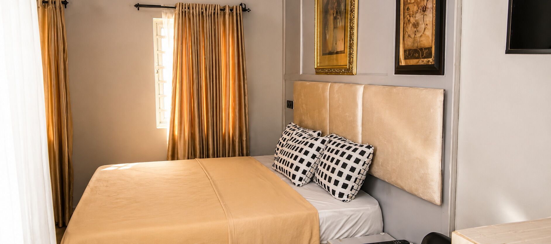 Una habitación de hotel con cama, cortinas, cabecero y dos cuadros en la pared.