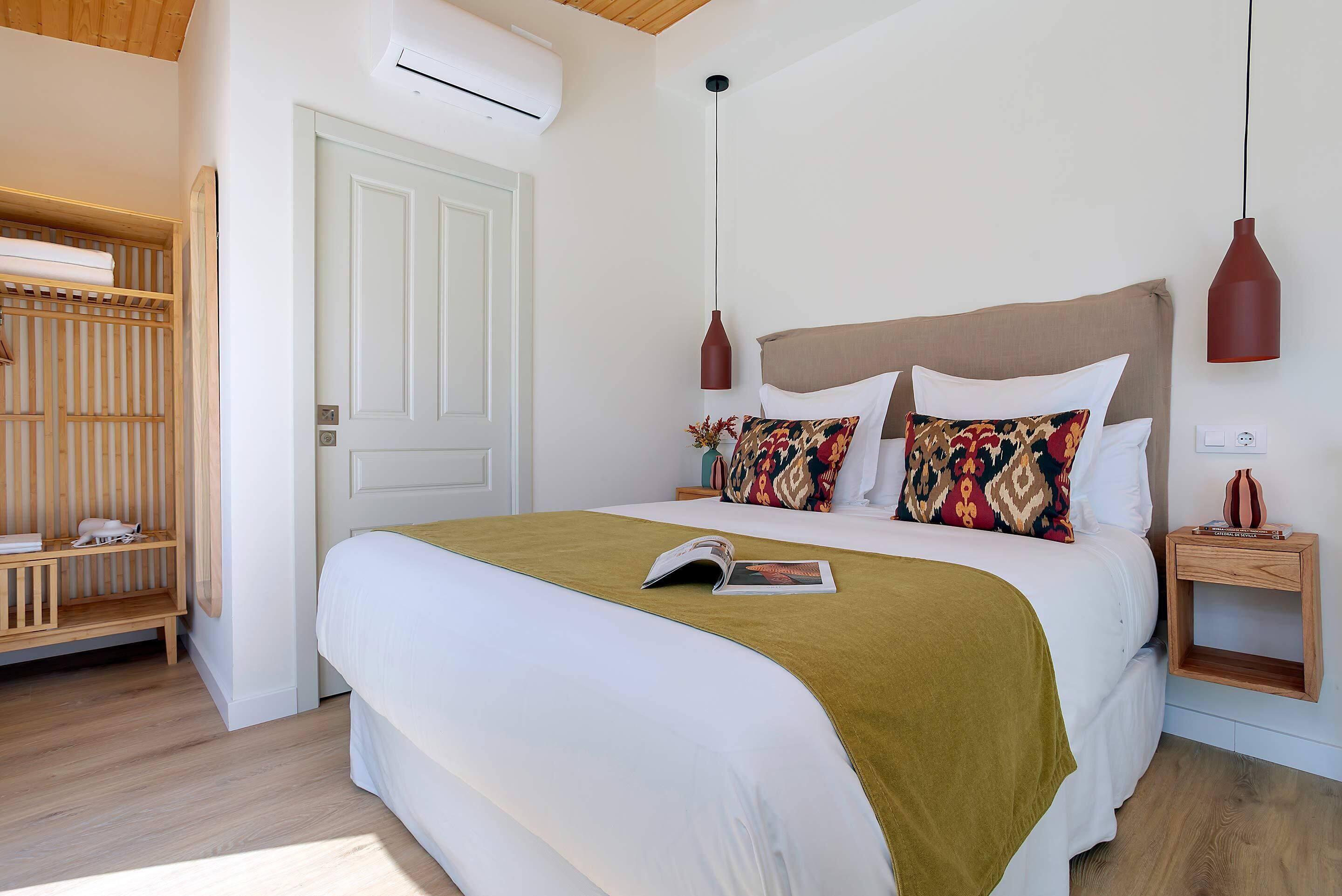 A neatly made bed with a green throw, a wooden bedside table with a book, and a hanging pendant light.