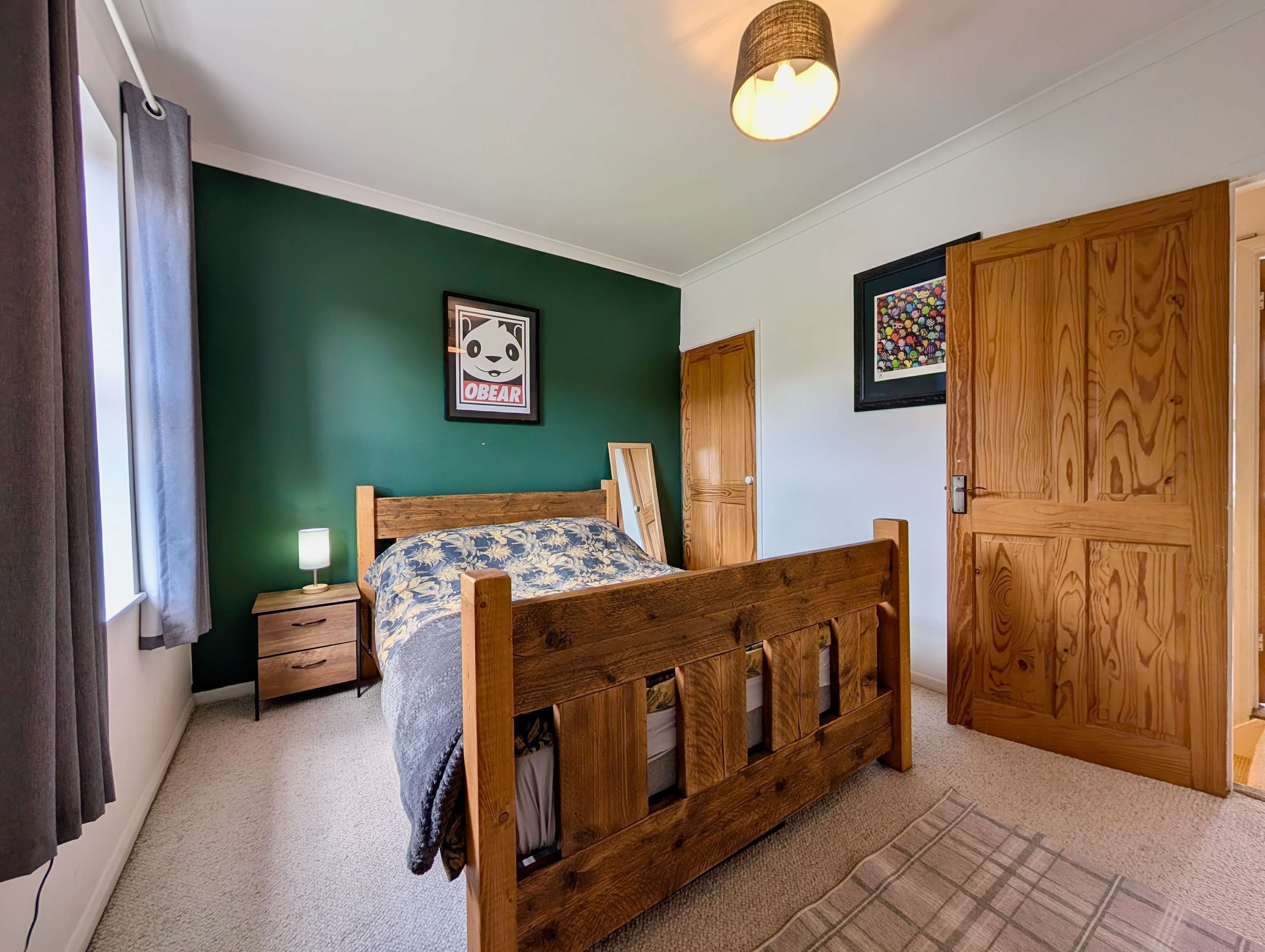 Quarto com cama de madeira, mesa de cabeceira, luminária, pôster com tema de panda, guarda-roupa de madeira e porta com arte colorida.