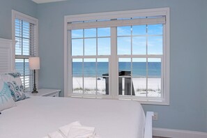 Een slaapkamer met een groot raam dat uitkijkt op een strand, een bed met witte lakens en een nachtkastje met een lamp.