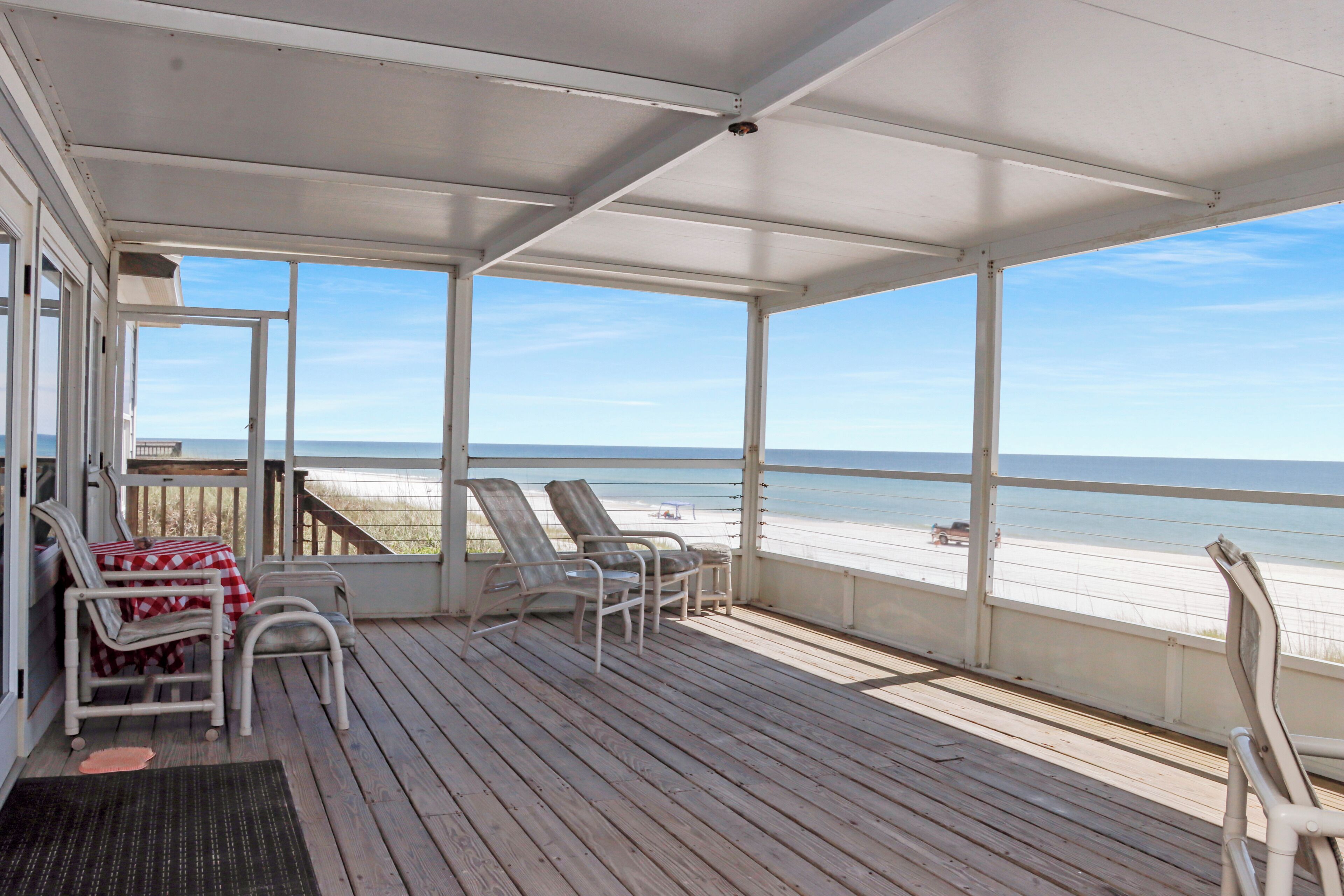 Um deck coberto com vista para a praia, cadeiras brancas e uma mesa com toalha quadriculada.