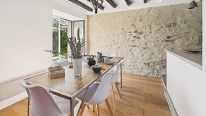 Uma área de refeições com mesa de madeira, cadeiras brancas, um vaso com plantas e uma parede de pedra.