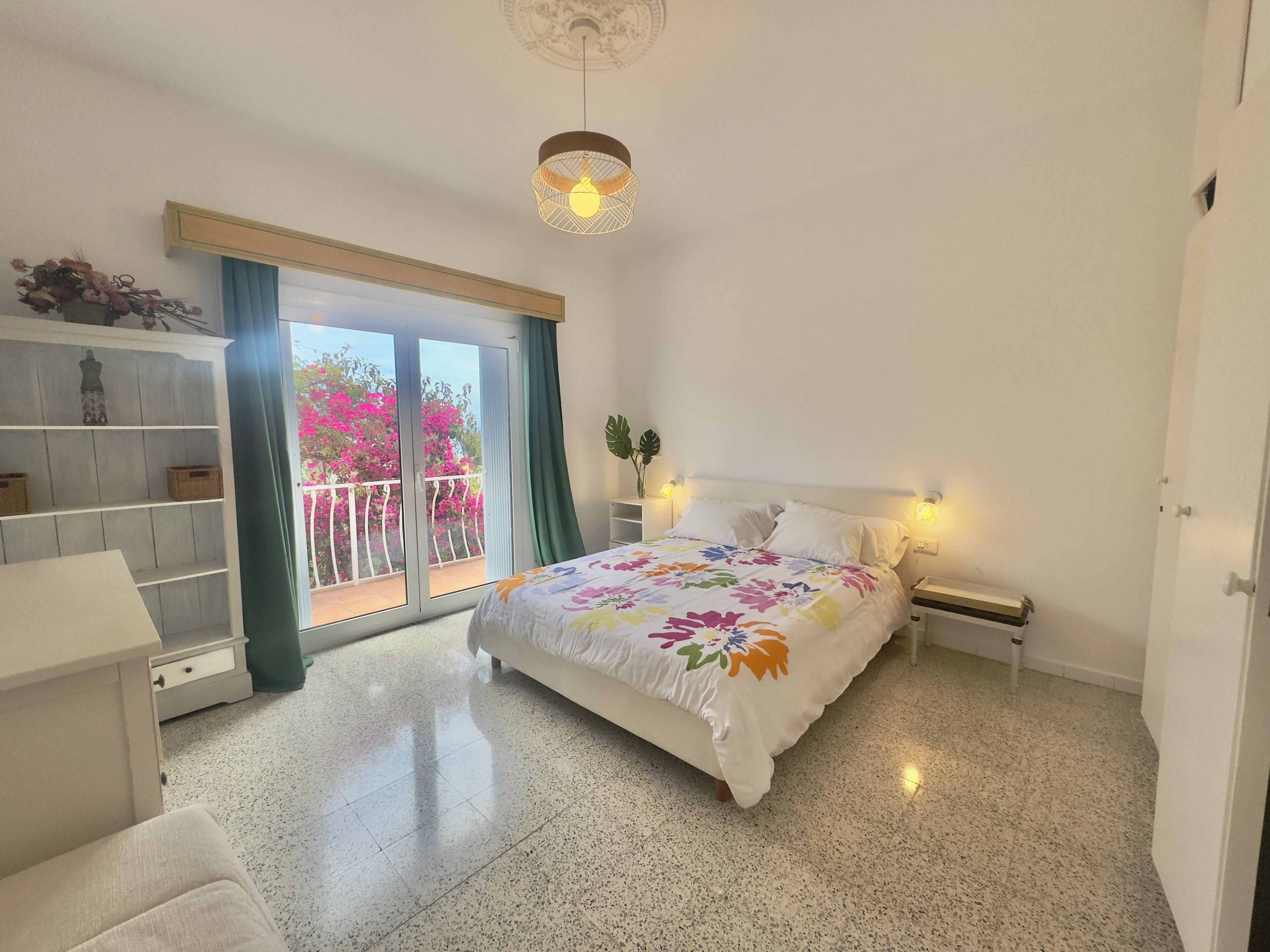 Een slaapkamer met een bed, een nachtkastje, een lamp, een plank met decoratieve objecten, en uitzicht op een balkon met roze bloemen.