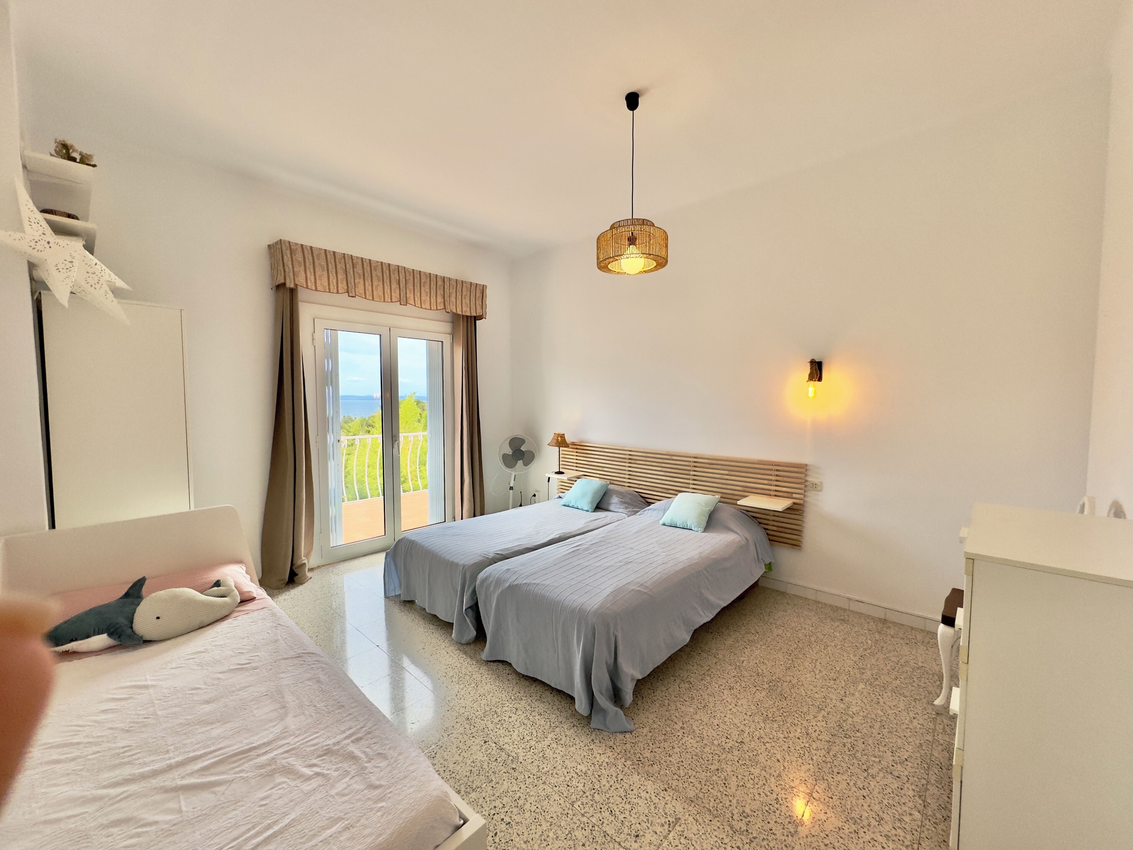 A bedroom with a bed, a bedside table, a lamp, a fan, and a view of the outdoors.