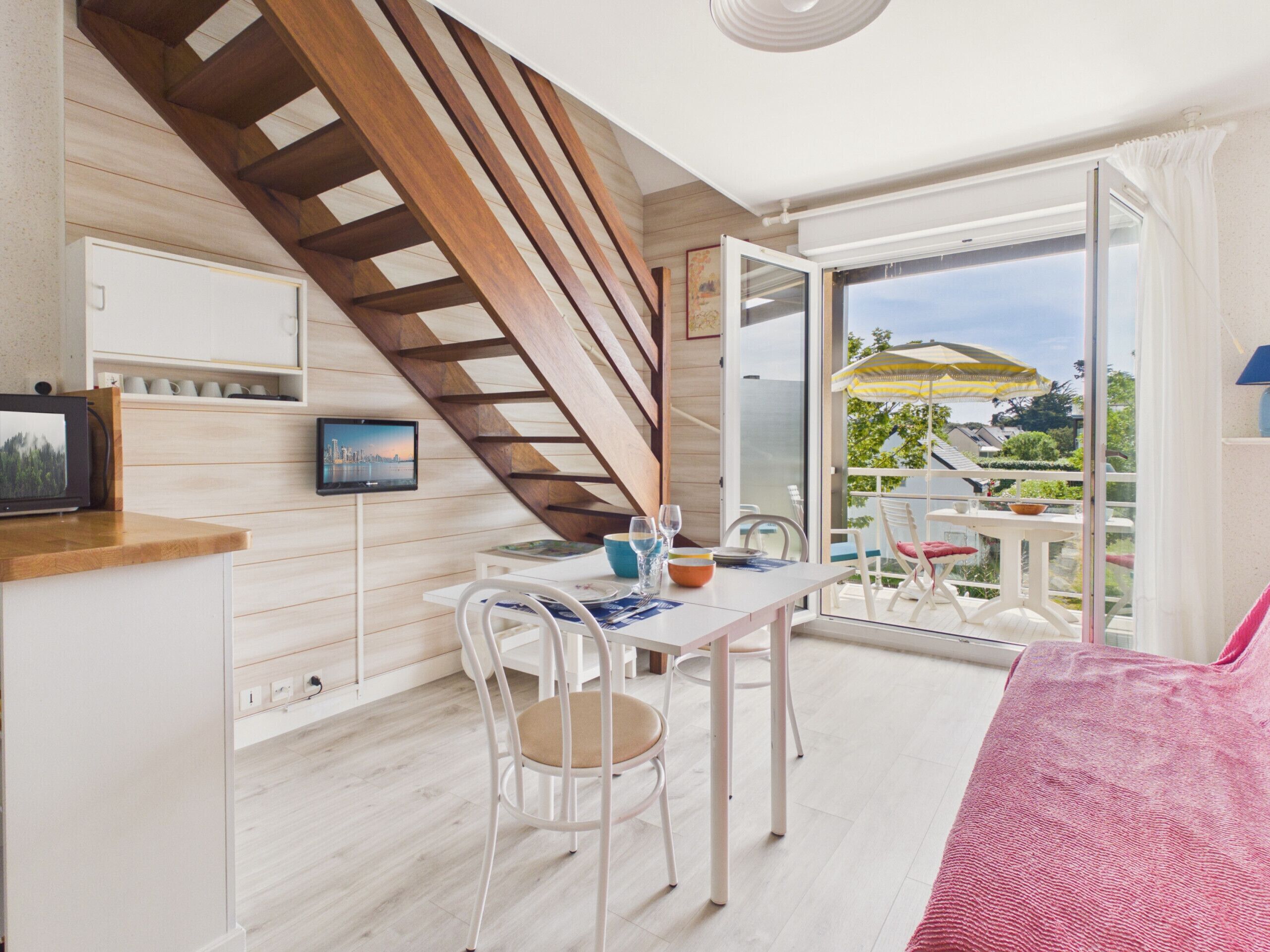 A modern room with a wooden staircase, a dining area with a table and chairs, a TV mounted on the wall, and a sliding glass door leading to an outdoor area with a table and umbrella.
