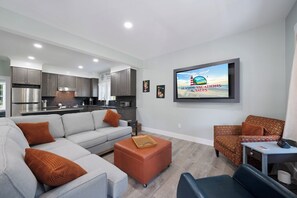A modern living room with a grey sofa, a brown ottoman, a patterned armchair, and a flat-screen TV mounted on the wall.