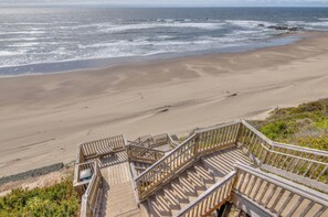 Eine Holztreppe führt zu einem sandigen Strand, im Hintergrund das Meer mit Wellen.