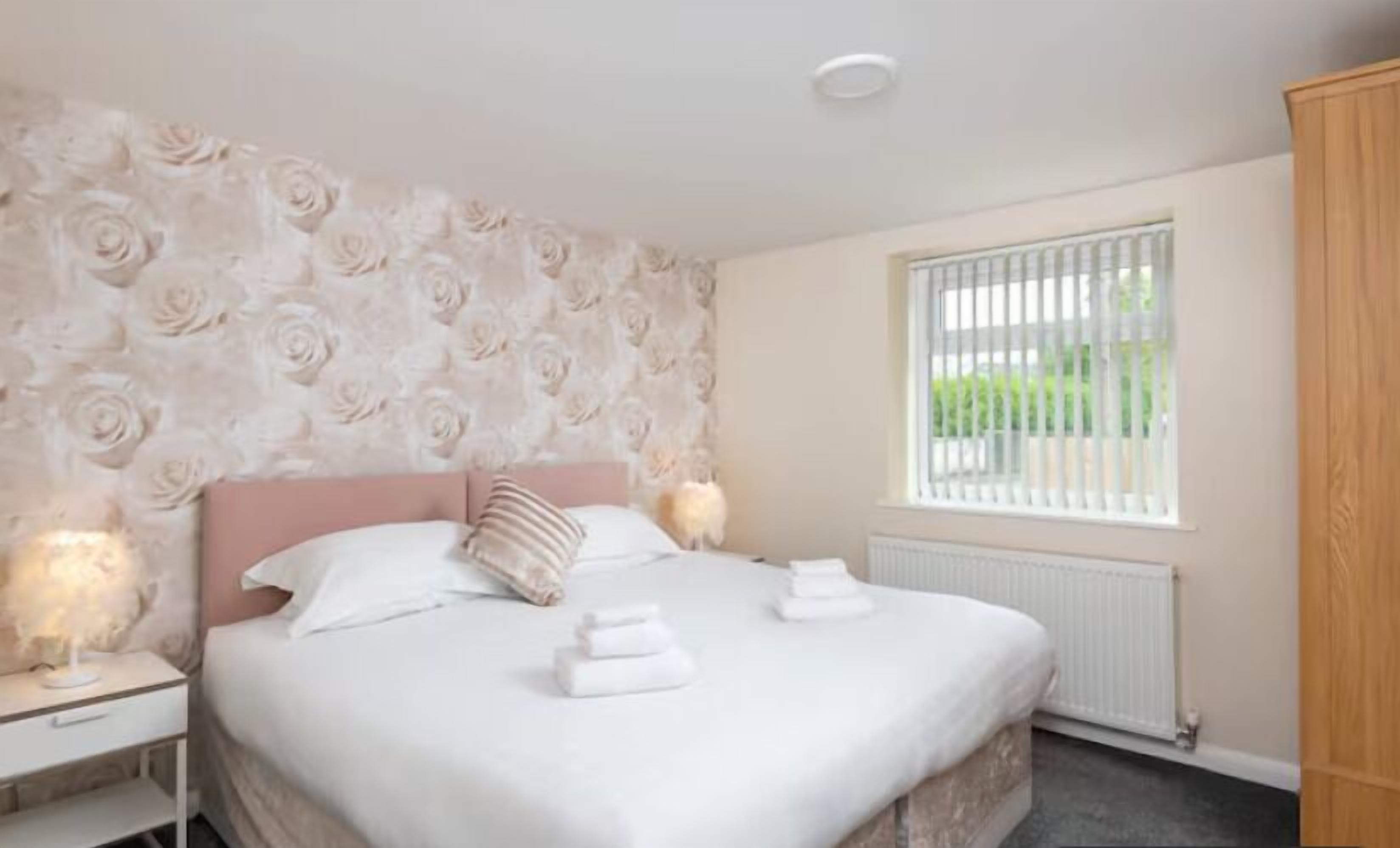 A bedroom with a large bed, a nightstand, a window with blinds, and a radiator.