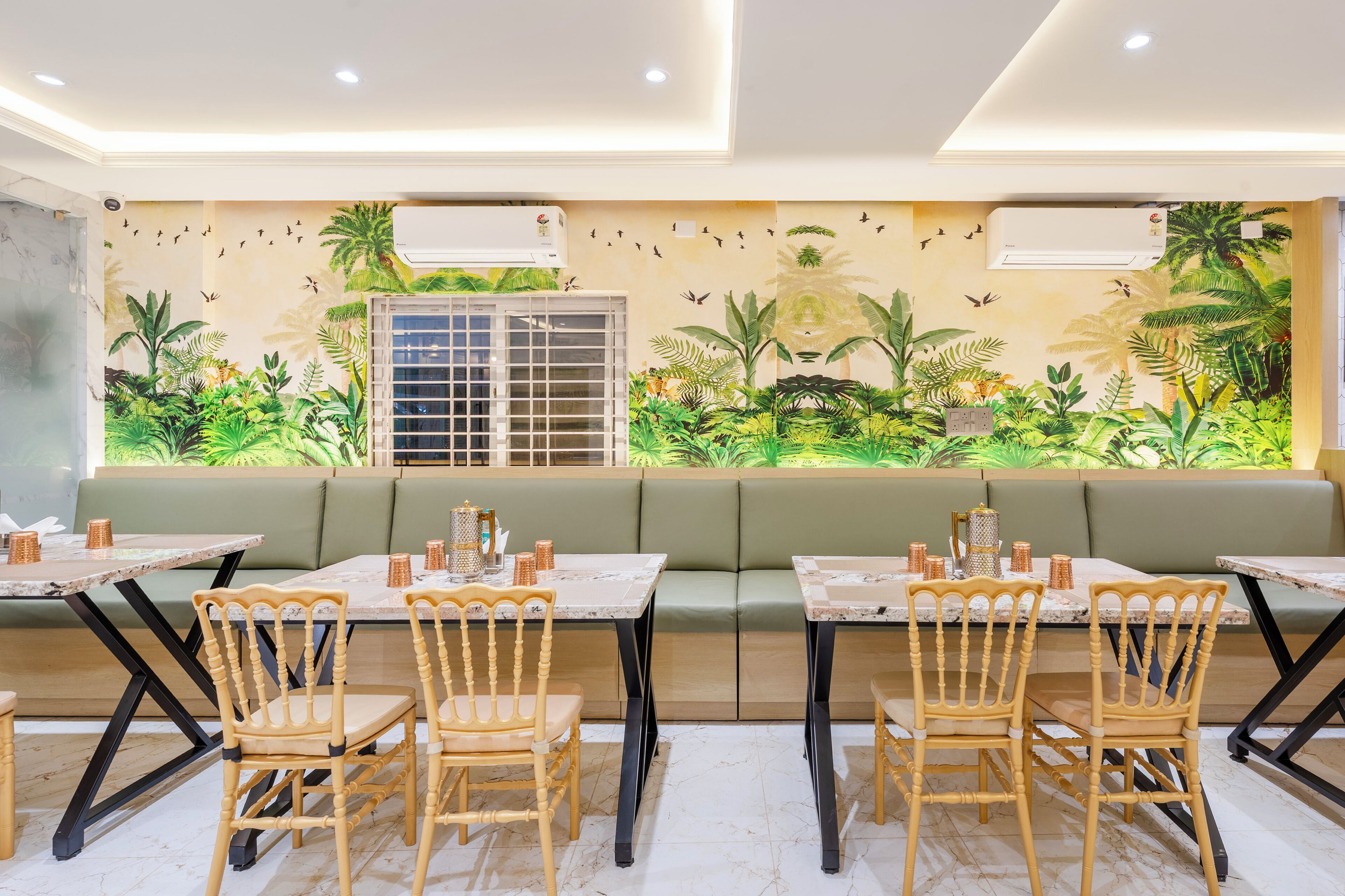 Un restaurant doté d’un coin salon avec des bancs rembourrés de tissu vert, des chaises en bois, des tables en marbre et une fresque murale représentant un paysage tropical.