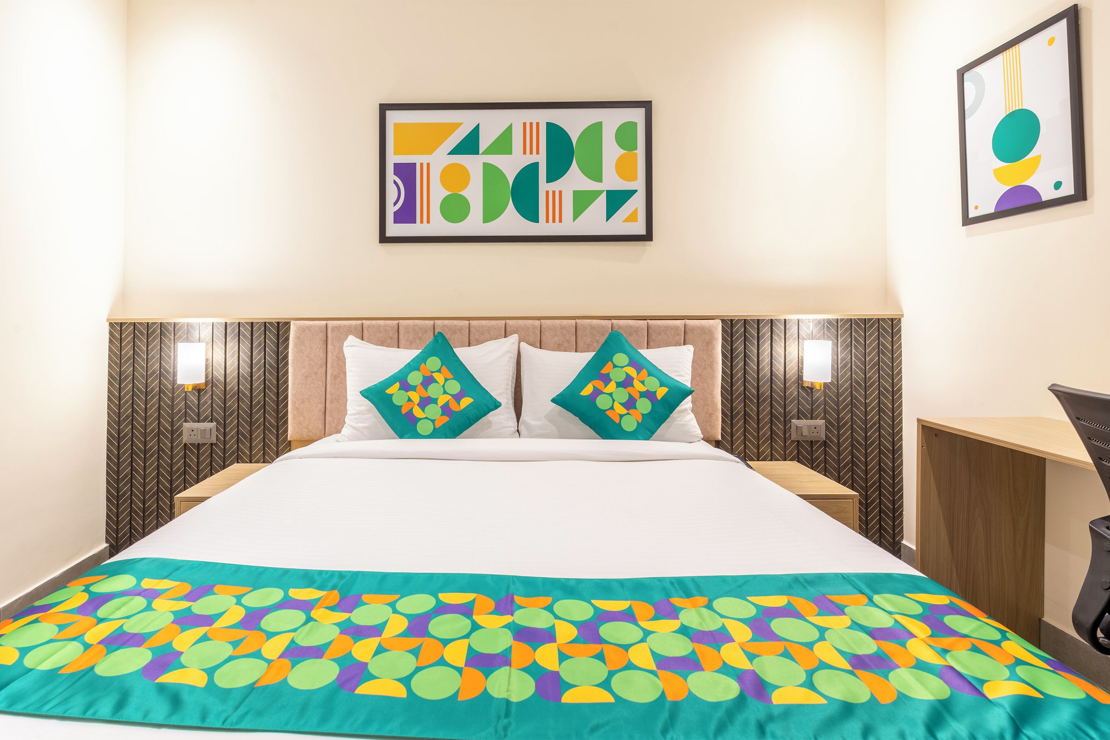 A hotel room with a bed, two pillows, a colorful blanket, a wall-mounted artwork, and a nightstand.