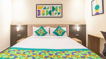 Una habitación de hotel con una cama, dos almohadas, una manta colorida, una obra de arte en la pared y una mesita de noche.