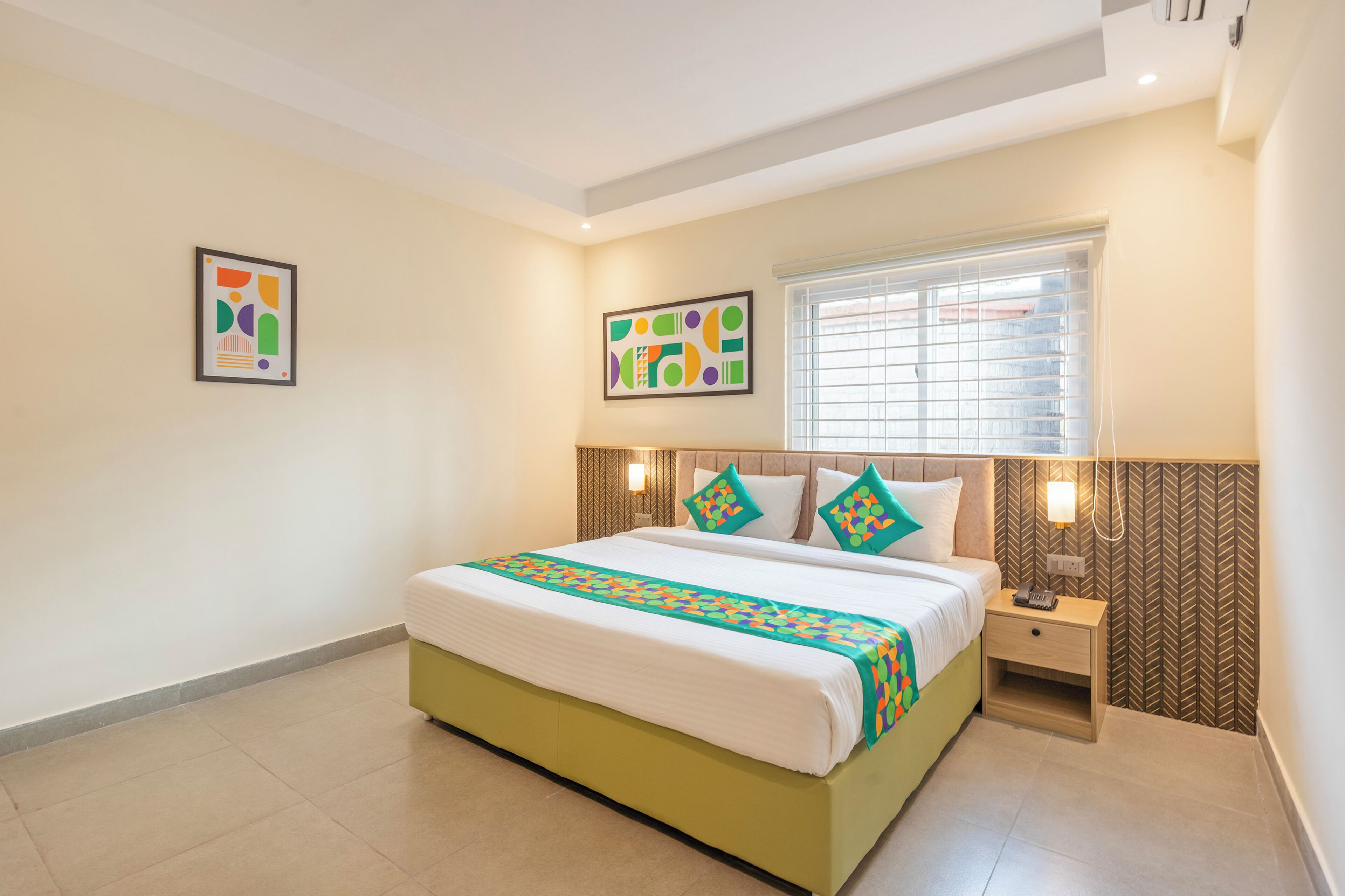 A hotel room with a bed, bedside table, wall art, and a window with blinds.