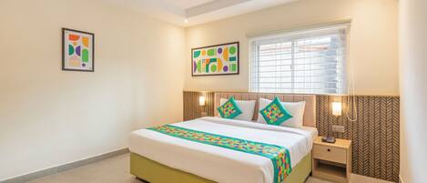 A hotel room with a bed, bedside table, wall art, and a window with blinds.