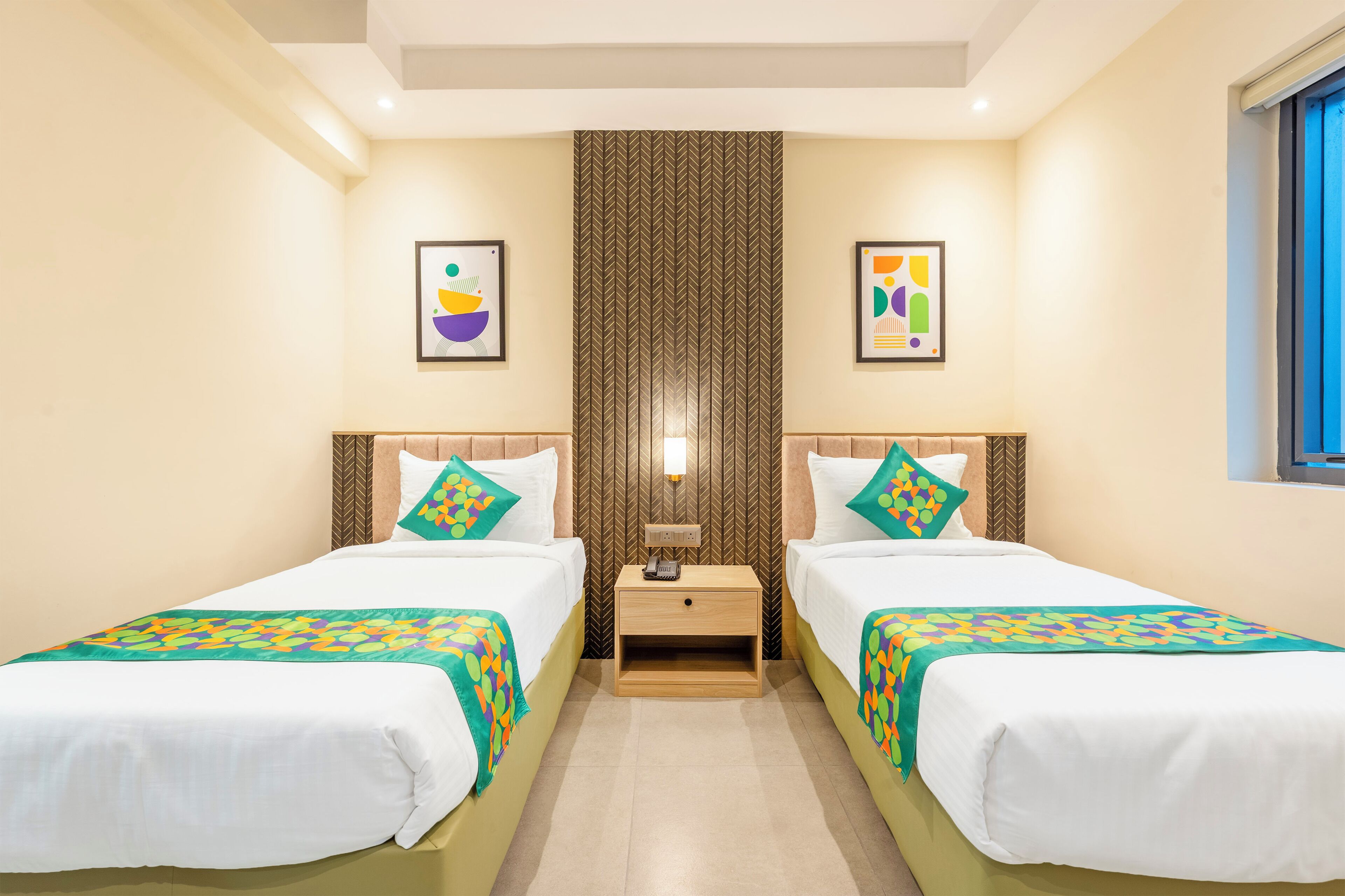 Una habitación de hotel con dos camas, una mesita de noche, una lámpara de pared y dos obras de arte enmarcadas.