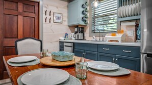 A dining table set with plates, a bowl, and glasses, with a clock and kitchen cabinets in the background. - 'Lil' Gem'-restored 1960's bungalow (Mentone)