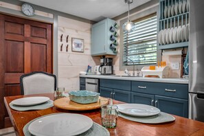 Une table à manger dressée avec des assiettes, un bol et des verres, avec une horloge et des meubles de cuisine en arrière-plan.