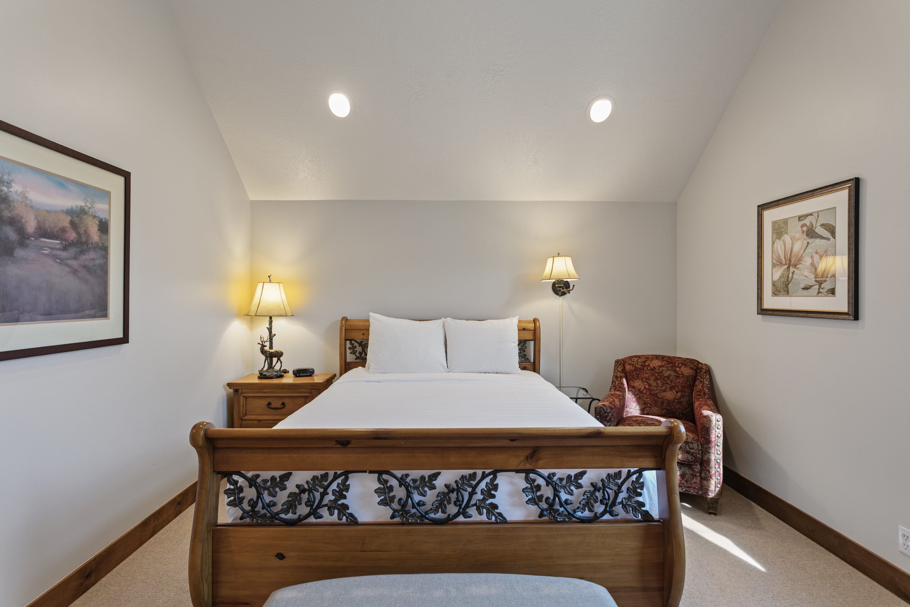 A bedroom with a wooden bed, two bedside lamps, a chair, and framed artwork on the walls.