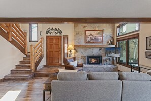 A living room with a fireplace, a stone wall, a wooden staircase, and a large window. - Deer Valley Townhome Walkable Location + Hot Tub 3 Bedroom Condo (Park City)