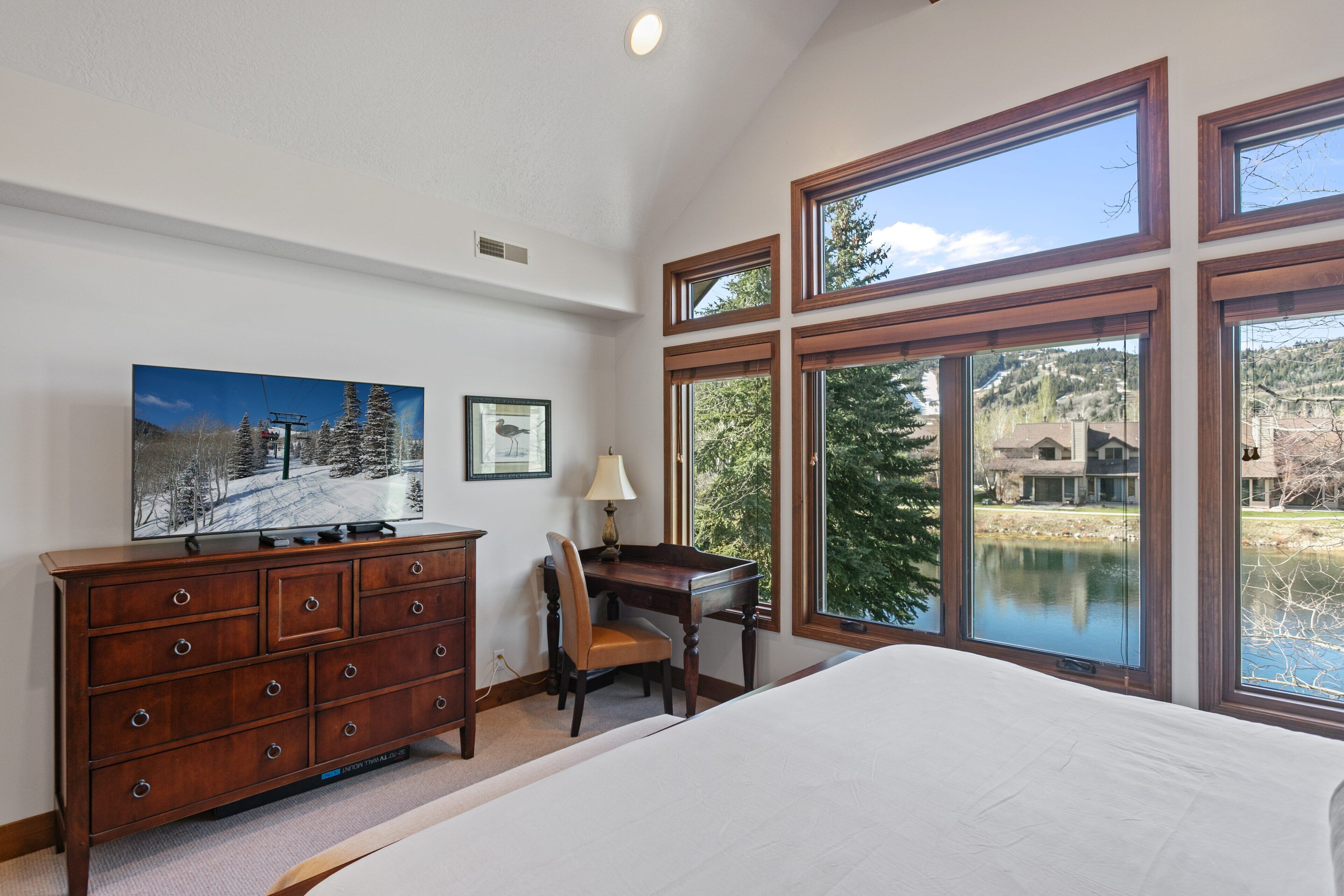Ein Schlafzimmer mit einem großen Bett, einer Holzkommode, einem Schreibtisch und einem Sessel. Mehrere Fenster bieten Blick auf einen See und Bäume.