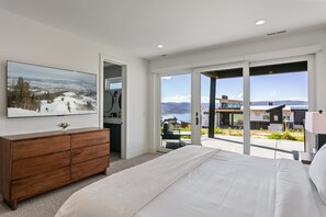 A bedroom with a large bed, a wooden dresser, a painting of a snowy landscape, and a sliding glass door leading to an outdoor area with a view of mountains and buildings. - Luxury Deer Valley East Village Home + Hot Tub 6br 6 Bedroom Home (Heber City)
