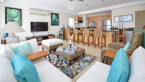 A modern living room with a sofa, coffee table, and a TV. There is a kitchen area in the background with bar stools and a dining table. - Palm Beach 610 2 Bedroom Apts (Hastings)