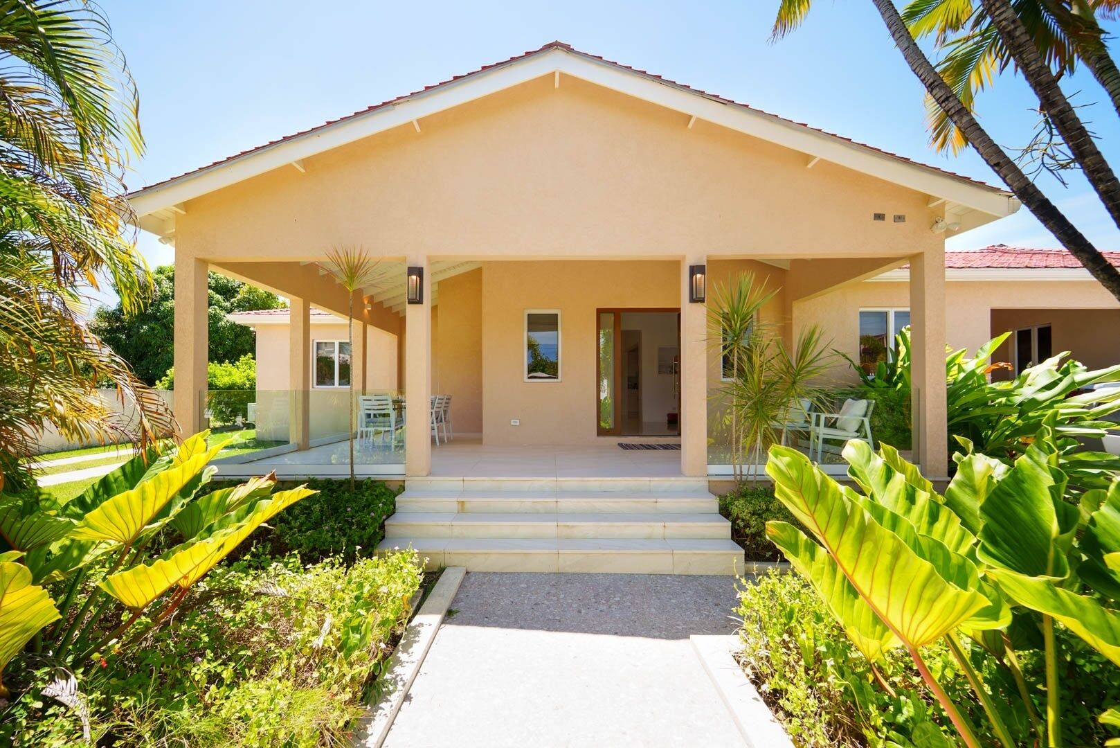 A modern building with a covered entrance, steps leading up to it, and a garden with tropical plants.
