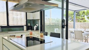 A modern kitchen with a central island, a dining area with white chairs, and a view of the outdoors through large windows. - ALLONBAY beach front residence (Villajoyosa)