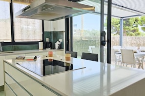 A modern kitchen with a central island, a dining area with white chairs, and a view of the outdoors through large windows. - ALLONBAY beach front residence (Villajoyosa)