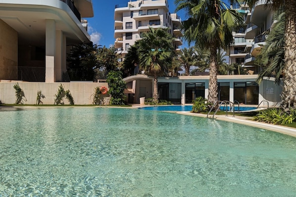 A swimming pool with a clear blue water, surrounded by palm trees and modern buildings. - ALLONBAY beach front residence (Villajoyosa)