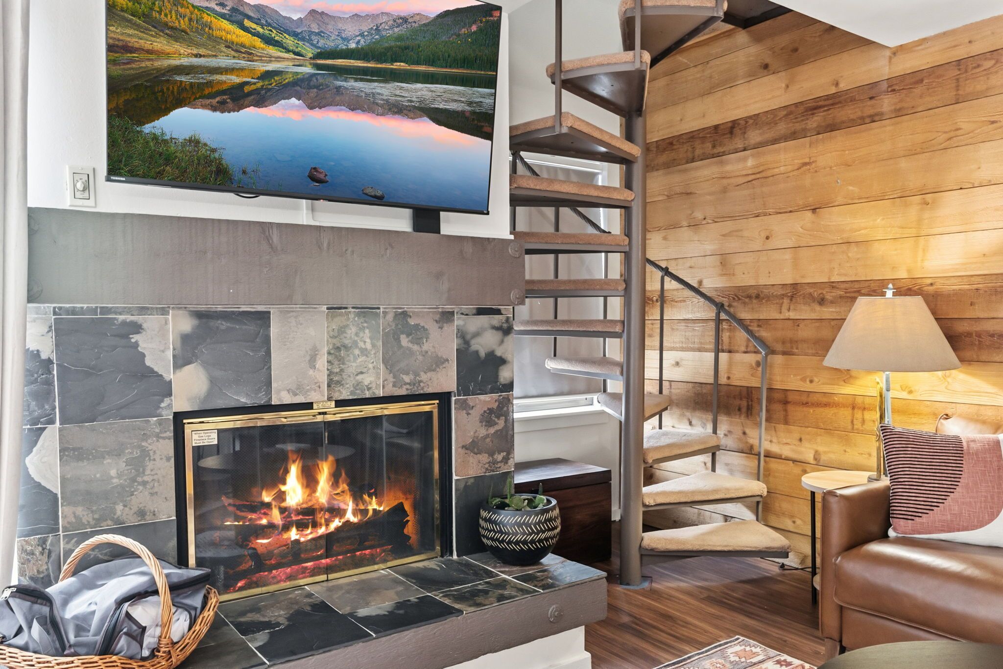 A modern living room with a fireplace, a television mounted above it, and a wooden spiral staircase.