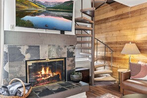 A modern living room with a fireplace, a television mounted above it, and a wooden spiral staircase. - Keystone Lakeside Condo | Outdoor Pool, Shuttle, and Mountain Access (Keystone)