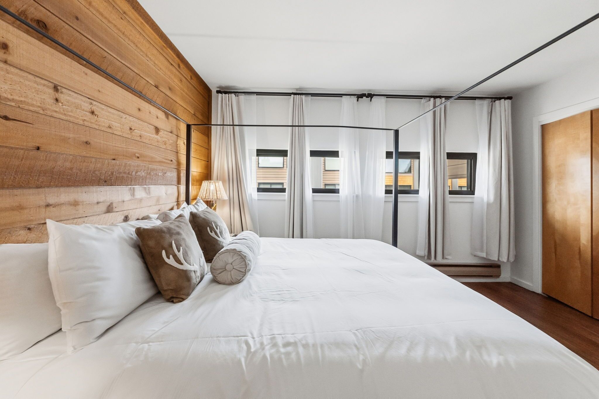 A bedroom with a wooden headboard, a large bed with white bedding, a window with curtains, and a view of buildings outside.