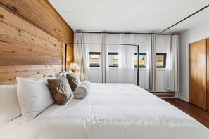 A bedroom with a wooden headboard, a large bed with white bedding, a window with curtains, and a view of buildings outside. - Keystone Lakeside Condo | Outdoor Pool, Shuttle, and Mountain Access (Keystone)