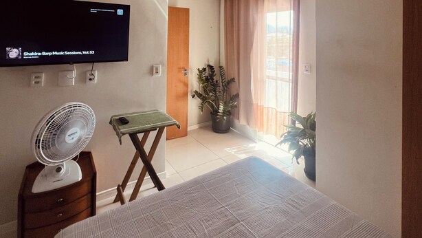 A bedroom with a bed, a TV mounted on the wall, a standing fan, a small table, and a potted plant. - Studio apartment in Prainha de Muquiçaba, 5 minutes from Praia do Morro, excellent for couples. (Guarapari)