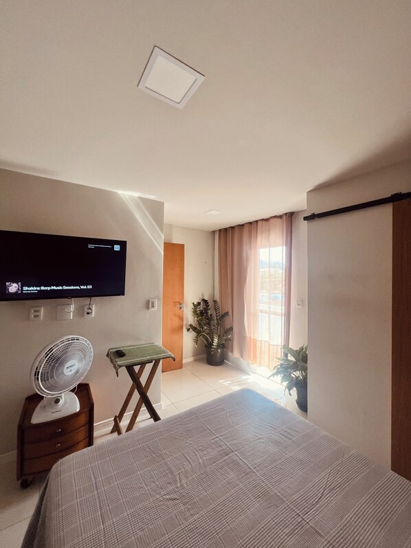 A bedroom with a bed, a TV mounted on the wall, a standing fan, a small table, and a potted plant. - Studio apartment in Prainha de Muquiçaba, 5 minutes from Praia do Morro, excellent for couples. (Guarapari)