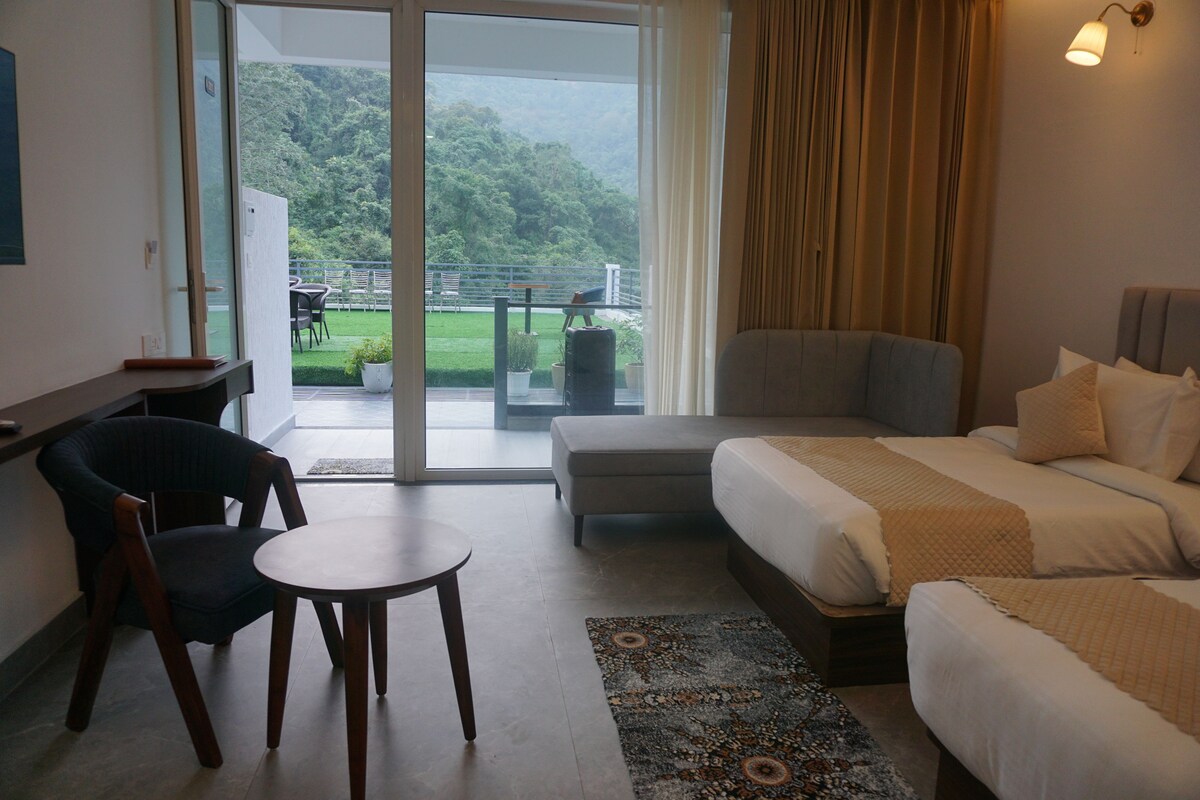 A hotel room with a bed, a chair, a small table, and a view of a green landscape through the window.