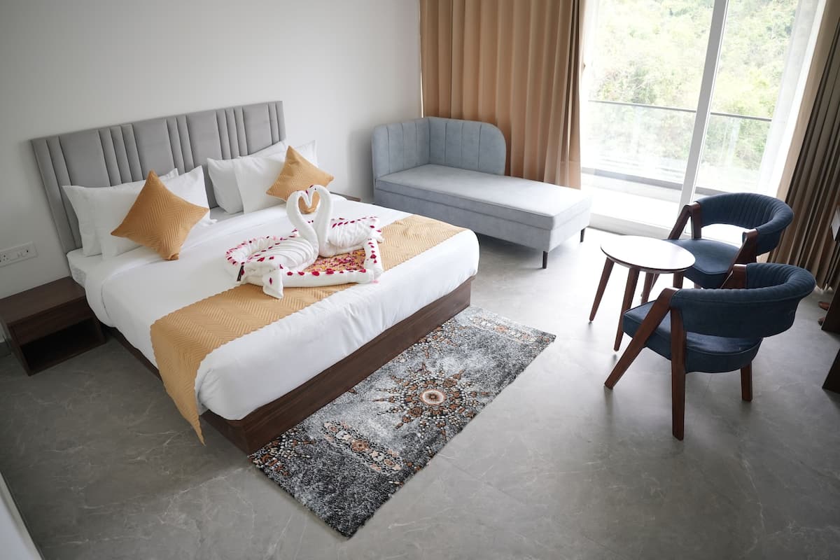 Une chambre d’hôtel avec un lit, un canapé gris, deux fauteuils bleus, une petite table ronde et une grande fenêtre avec des rideaux.