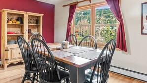 Un comedor con una mesa de madera, sillas negras, un armario de madera con vajilla y un ventanal con cortinas.