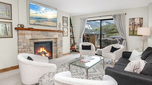 Un salon doté d’une cheminée en pierre, de meubles blancs et gris, d’une table basse en verre et d’une grande fenêtre offrant une vue sur les montagnes.