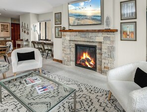 A living room with a fireplace, a dining area, and a large window with a painting. - Modern Condo in Avon, Walk to Gondola, SA513 (Avon)