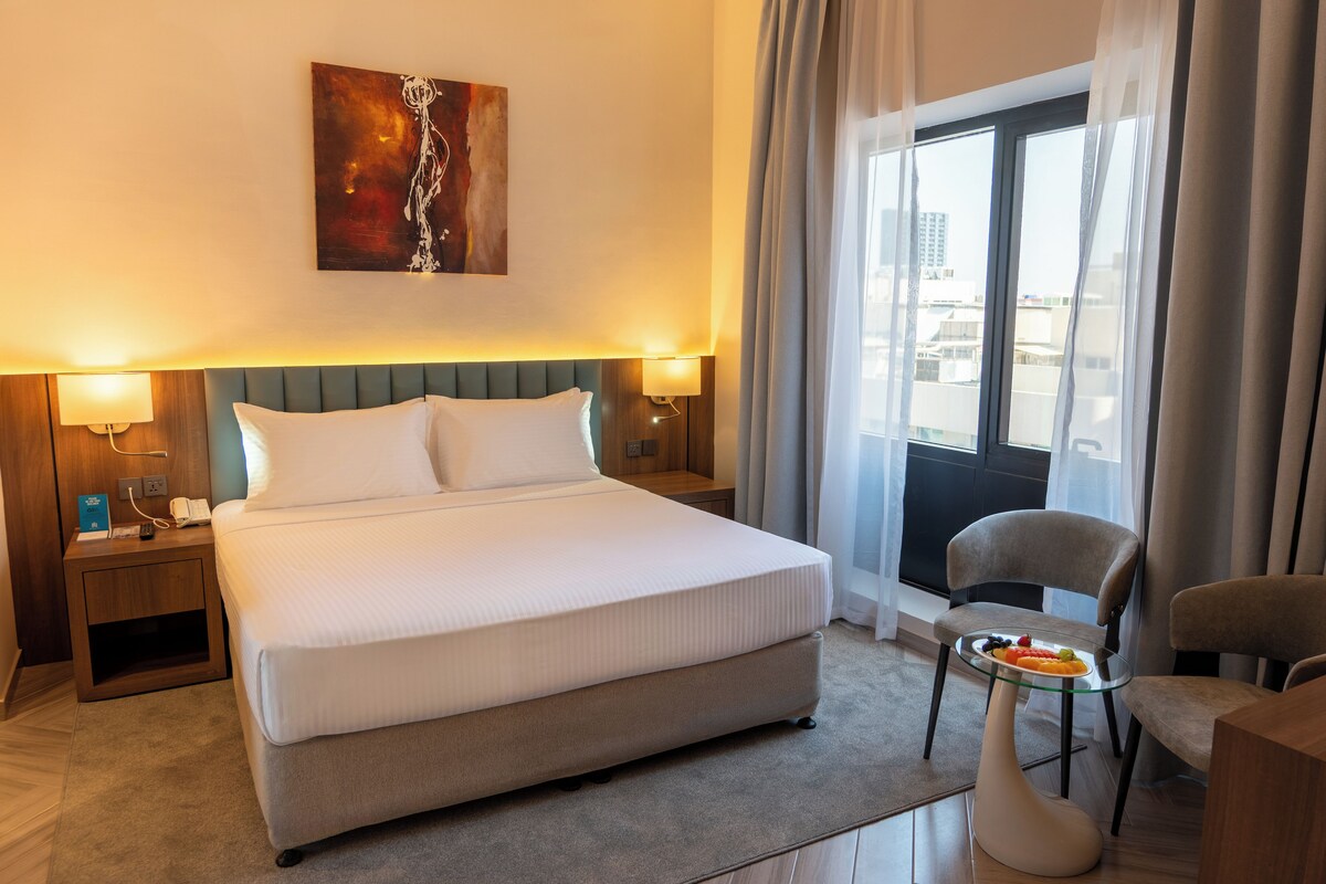 A hotel room with a large bed, bedside lamps, a small table with fruit, a chair, and a city view through the window.