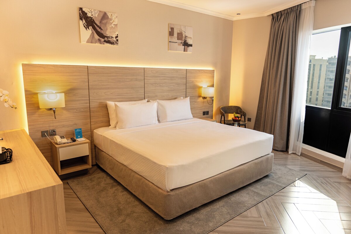A hotel room with a large bed, bedside lamps, a nightstand, and a view of the city through the window.