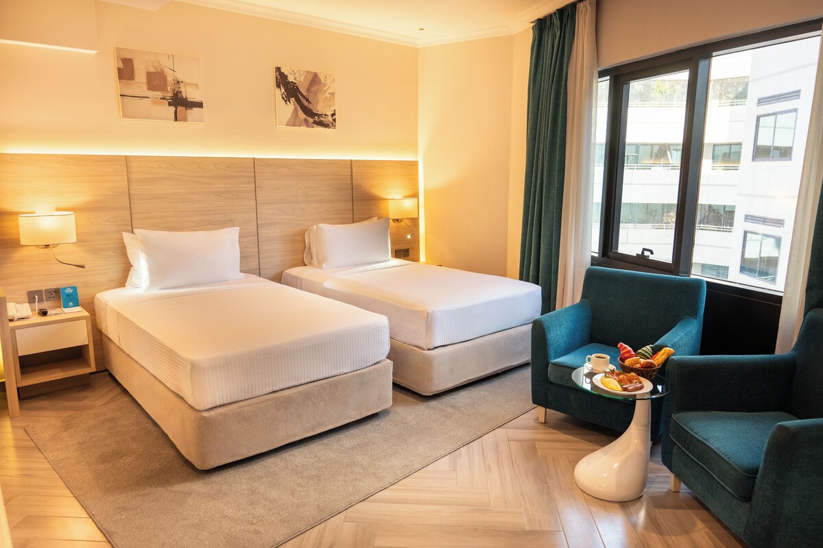A hotel room with two beds, a sitting area with a chair and a tray of breakfast, and artwork on the walls.